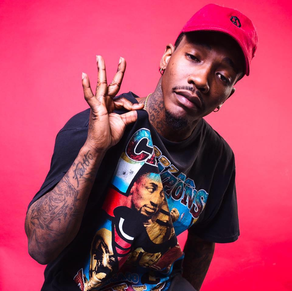 Indie rapper Dizzy Wright pulls up his shirt in front of a bright pink background.