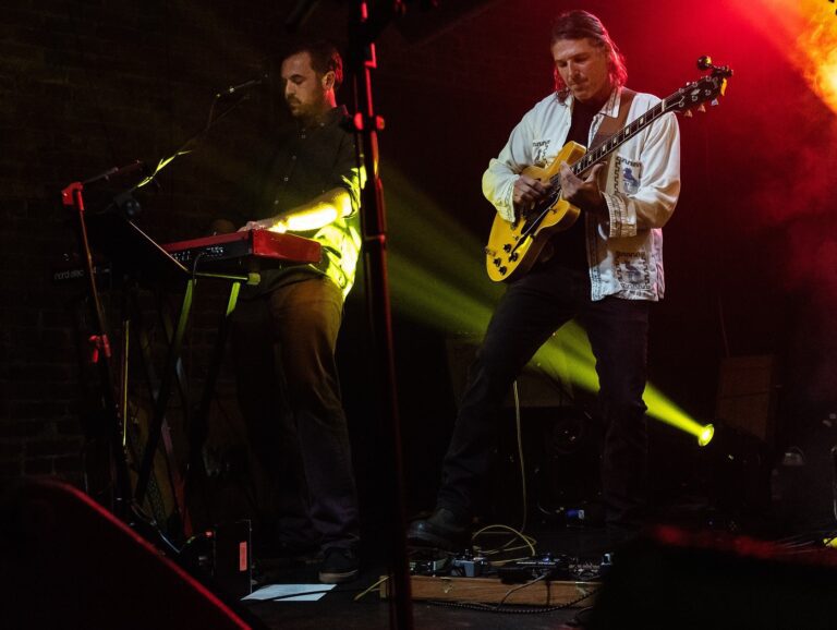 Bellingham musicians Aaron Guest, left, and Michael Iris play their respective instruments on stage with bright lights.