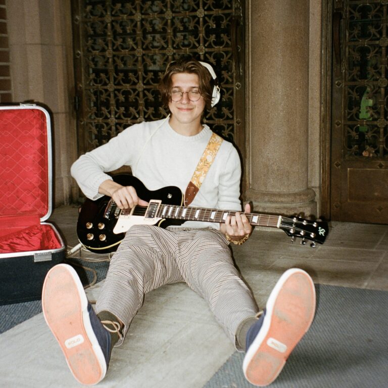 Austin Colwell sits next to his empty guitar case with the guitar strapped to him.