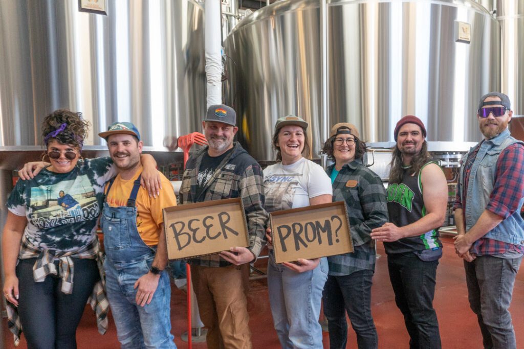 Beer Prom returns standing next to metal brewers with a cardboard sign asking "BEER PROM?"