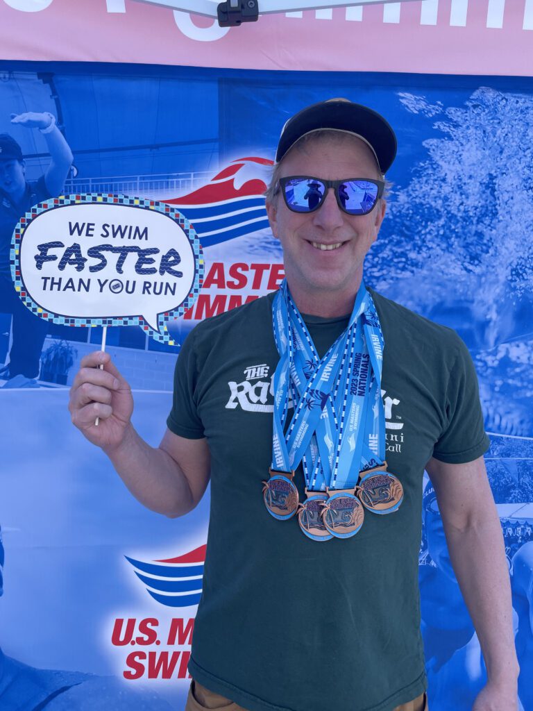 Brad Jones with four medals around his neck as he holds up a sign stating "We swim faster than you run."