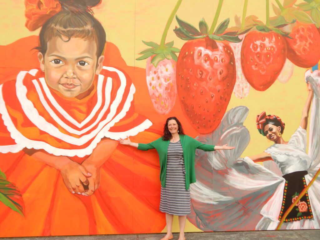 Mattix poses for a photo in between her murals with her arms spread wide.