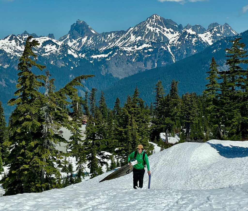 An outdoor adventurer climbs up a steep trail leaning onto a snowskate and holding onto a box of donuts close to him.