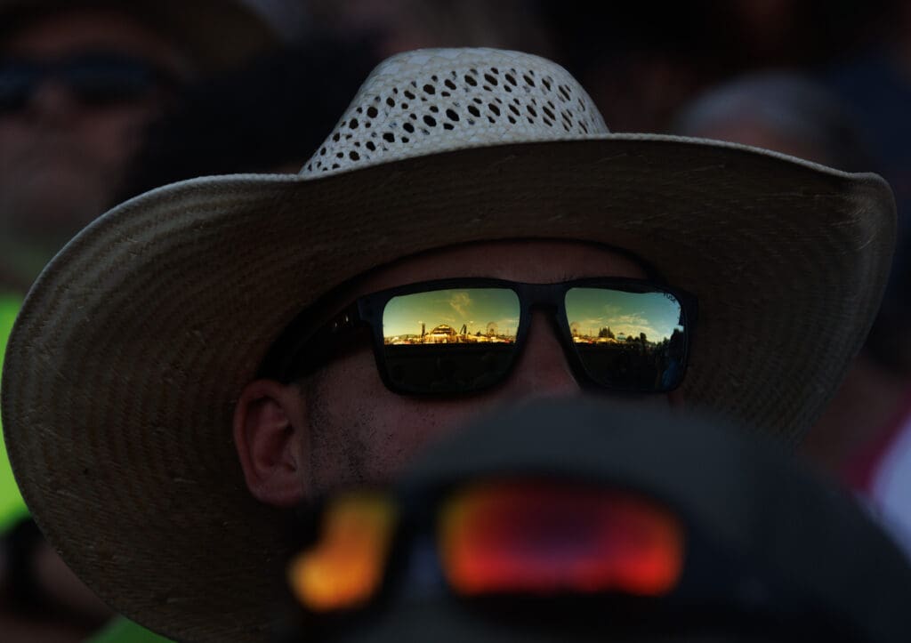 The fairgrounds can be seen in a reflection of a rodeo fan’s sunglasses.