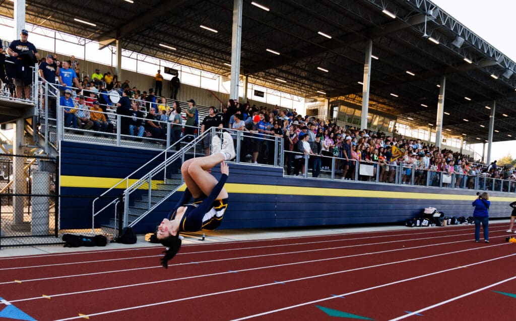 Cheerleader Giana Thompson practices a back tuck next to bleachers with many onlooking spectators.