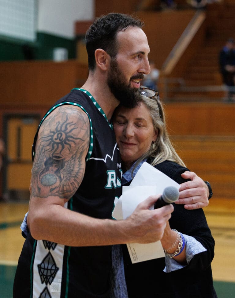 Tyler Amaya is hugged by Stacy Chapin while holding a microphone and a stack of papers.