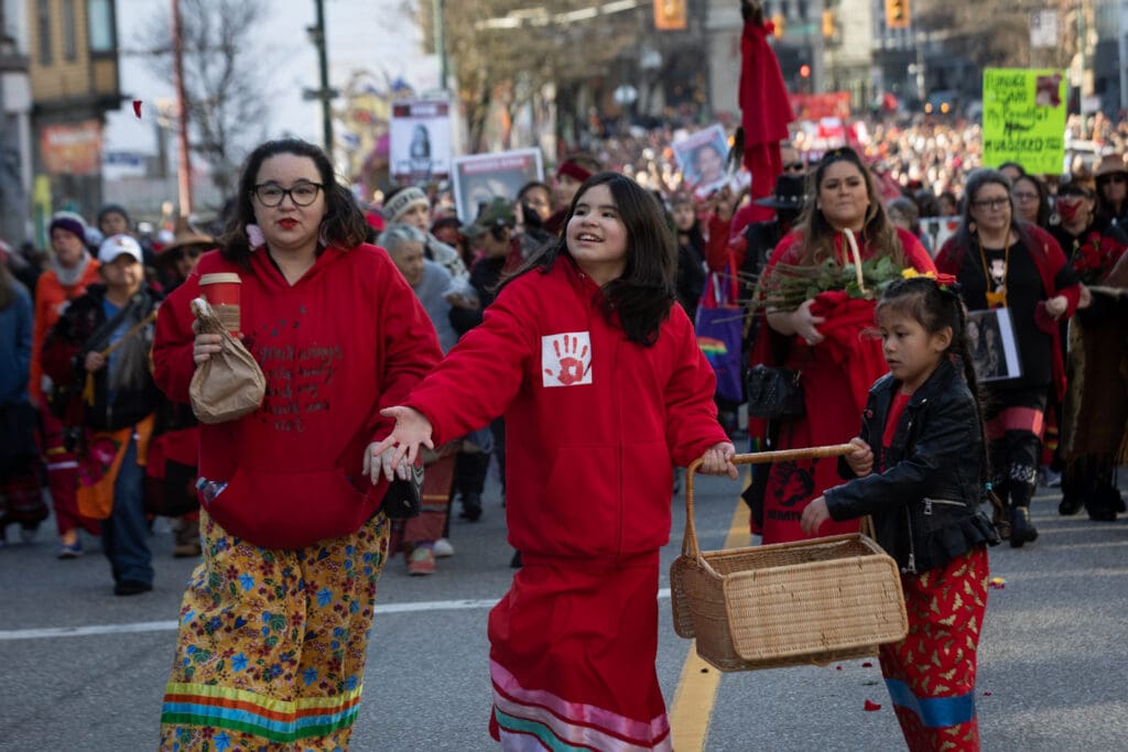 Girls wearing traditional ribbon skirts scatter flower petals at the front of the march.