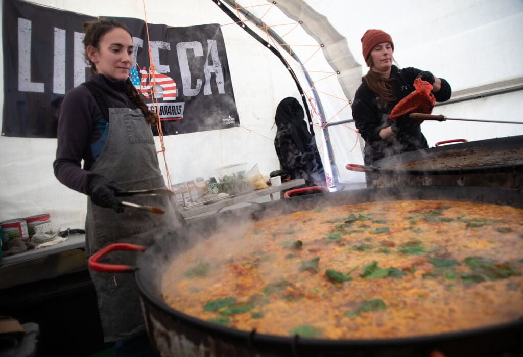 Dana Brownstein and Ona Osin cook paella in a large pan inside a white tent.