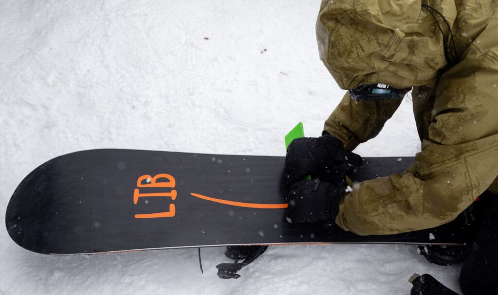 A snowboarder waxes their black snowboard with orange letters before their race.