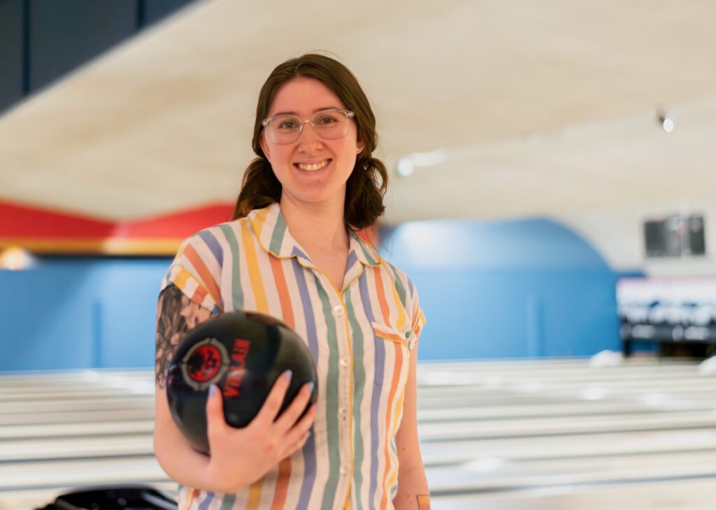 A young woman wearing glasses and a striped shirt stands in a bowling alley and poses for a photo while holding a blue and red bowling ball.