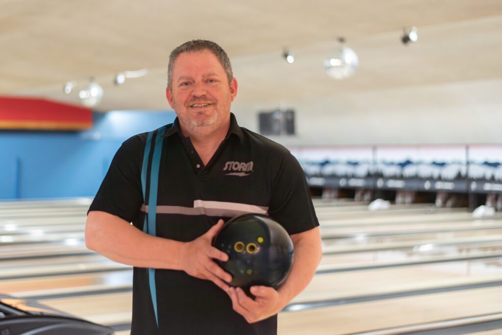 A man in a bowling shirt poses for a photo in a bowling alley while holding his bowling ball.