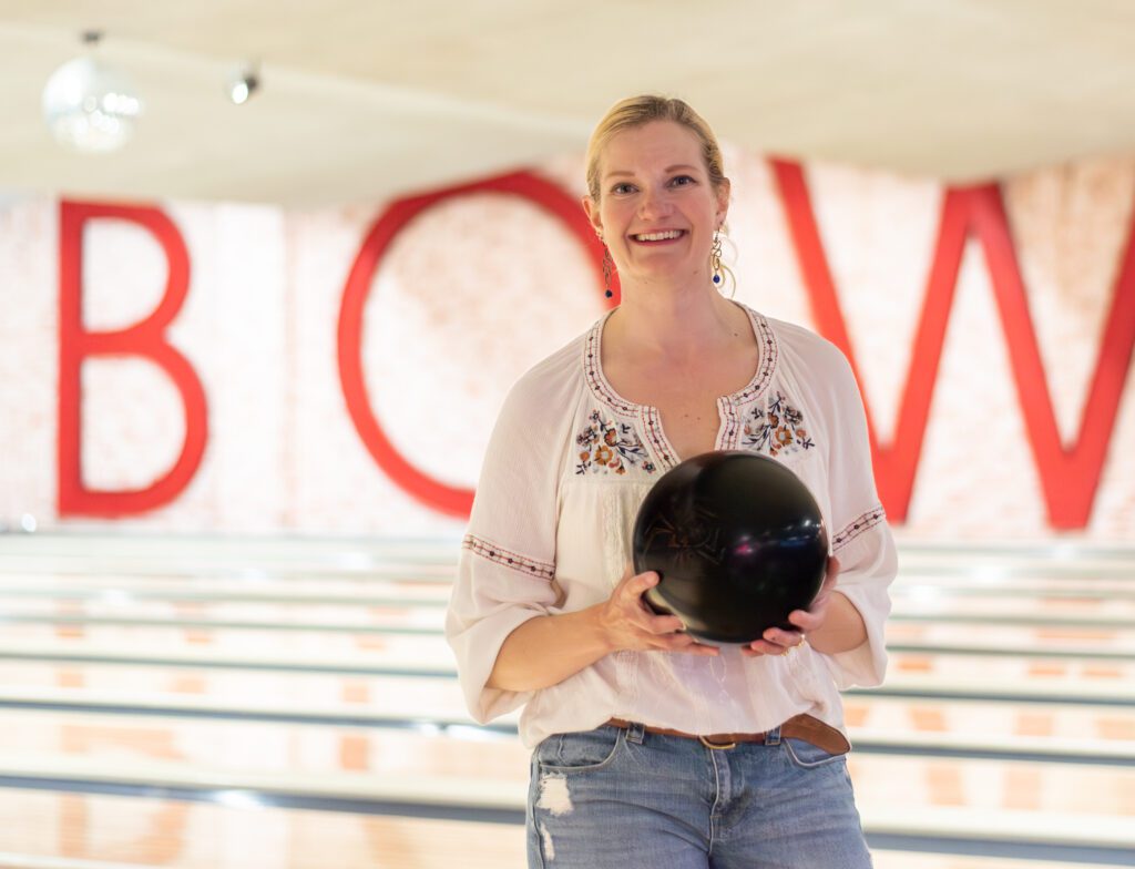 A woman in a white shirt holds a black bowling ball in a bowling alley. Behind her the word "BOWL" is in all red letters on a brick wall.