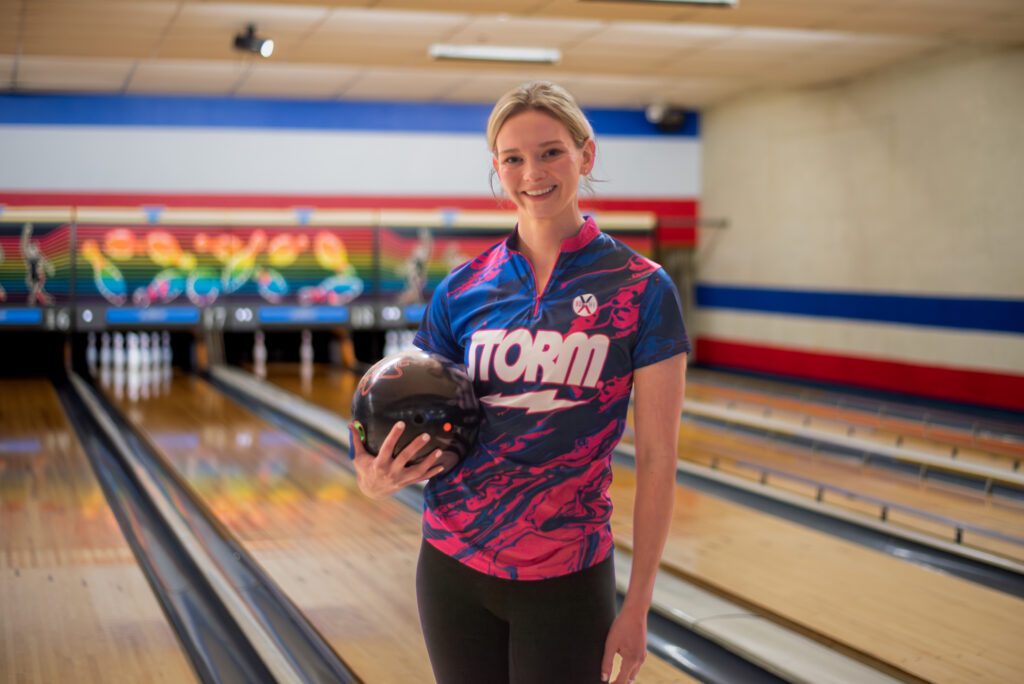 A young woman in a Storm bowling shirt holds a bowling ball while posing for photo in a bowling alley.