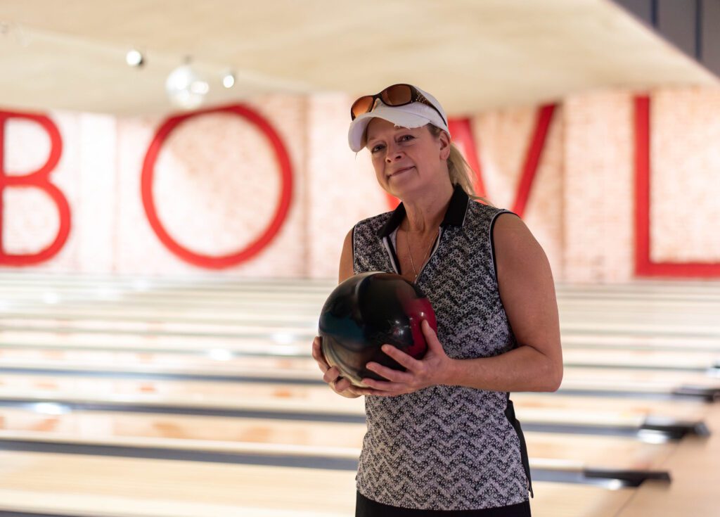 A woman in a white baseball hat poses for a photo while holding a bowling ball at a bowling alley. The word "BOWL" in red is behind her on a brick wall.