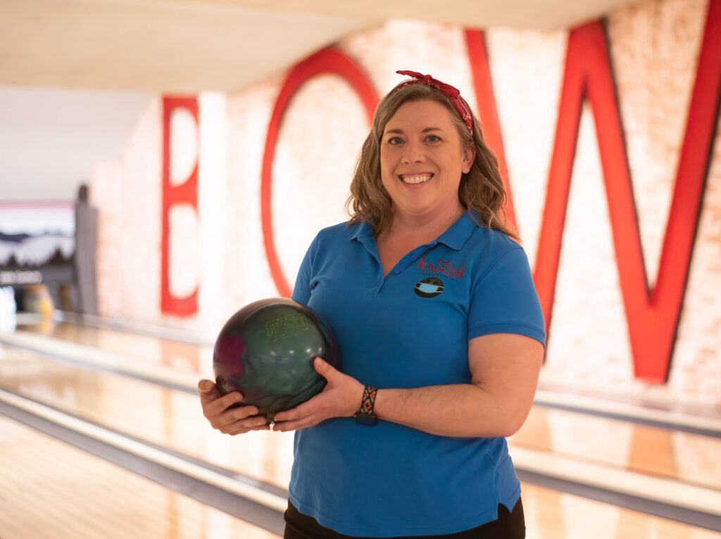 A woman in a blue shirt poses for a photo in a bowling alley while holding her bowling ball. "BOWL" in red letters is in the background on a brick wall.