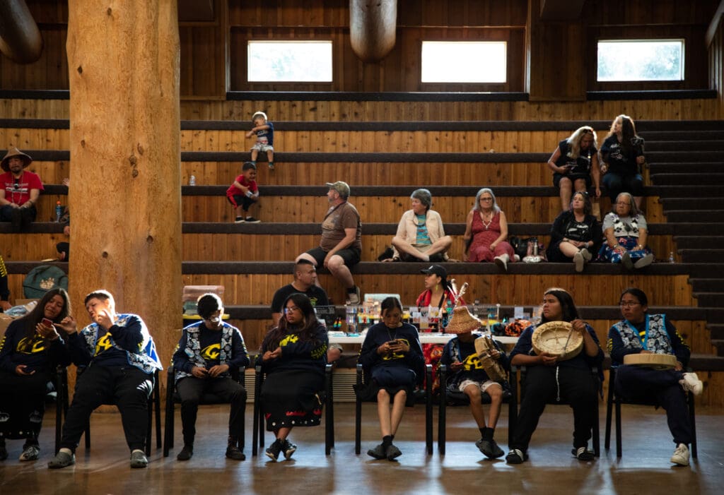 Visitors from the Chief Leschi School gather at the Wexliem Community Building as attendees sit at the wooden bleachers while vendors set up their booths behind performers.