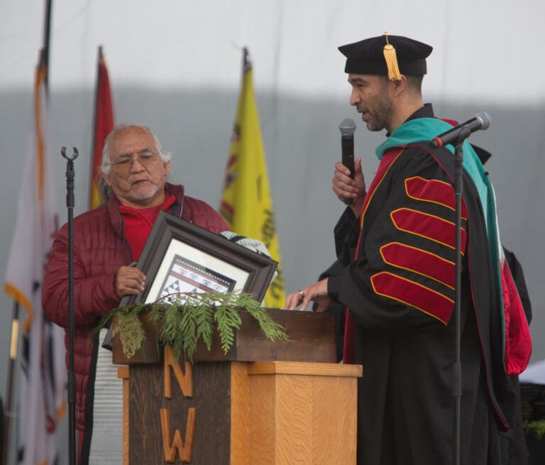 Justin Guillory, the president of Northwest Indian College and a descendent of the Nez Perce Tribe, honors Al Scott Johnny, a member of the Lummi Nation, with an honorary bachelor's degree as he delivers a speech with a microphone in hand.