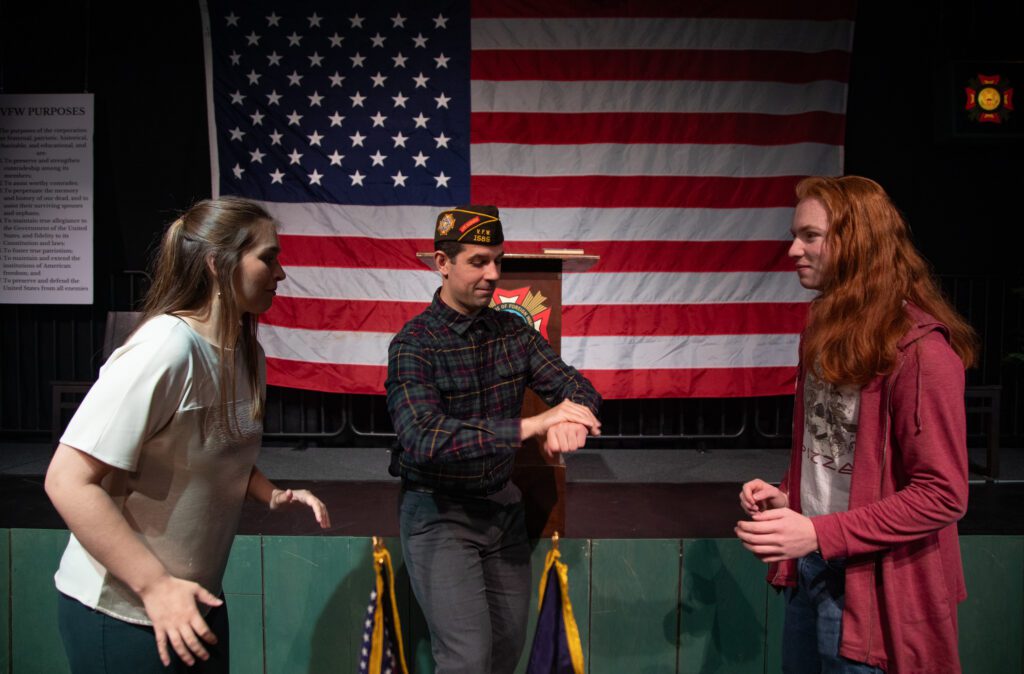 Adam St. John as the Legionnaire flips a coin to start a debate between actress Gabi Gilbride and Avery Horton in front of the stage.