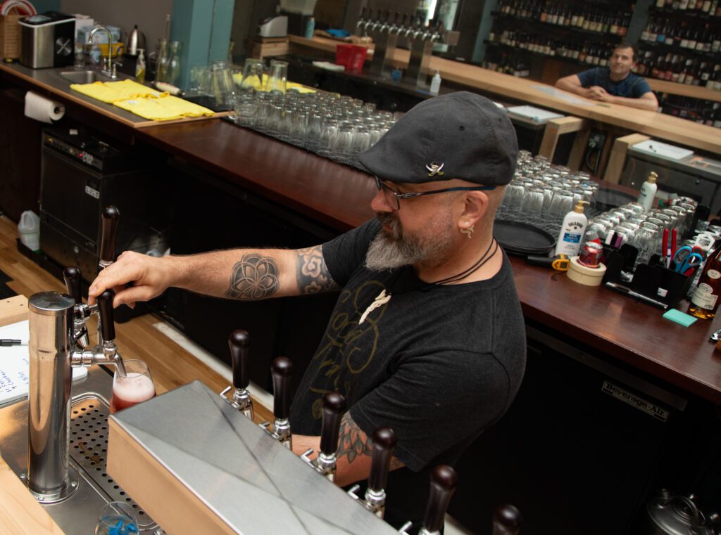 Joshua Brightware pulls a tap of beer for a customer with a mirror behind him showing a customer waiting at the bar counter.