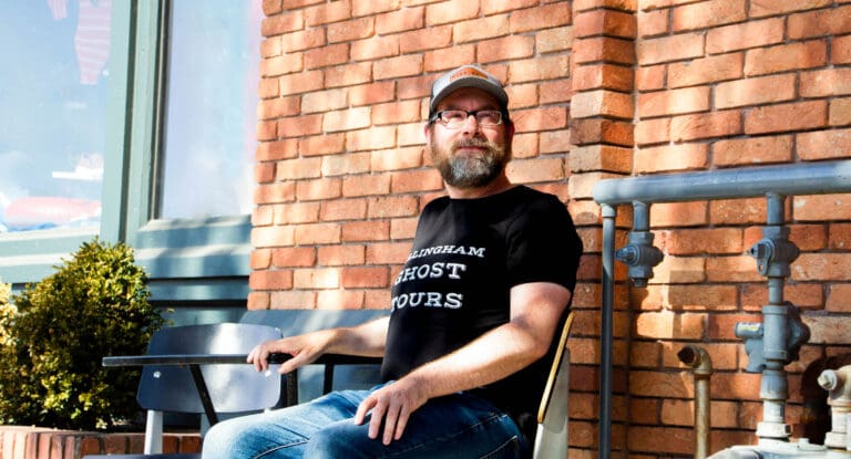 Charles Crooks, recently launching Bellingham Ghost Tours, sits on a bench with a brick wall behind him.