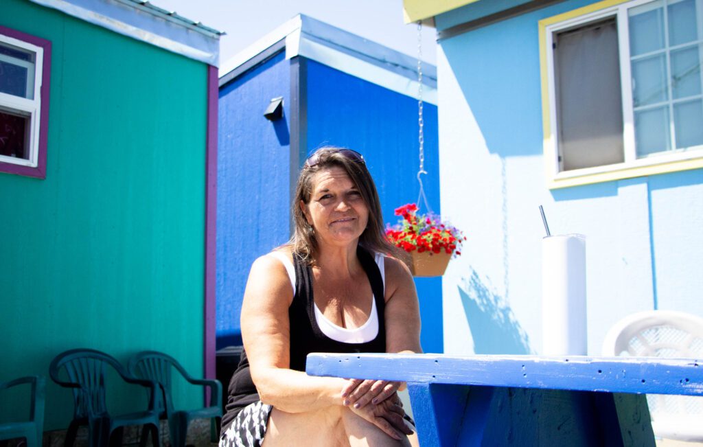 Tina Hayes, a resident of Unity Village, smiles for the camera while sitting at a blue table and surrounded by the colorful homes.