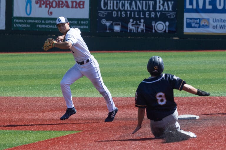 Bellingham Bells second baseman Cole Yoshida looks to throw as he twists while another player slides to the base with a trail of dirt.