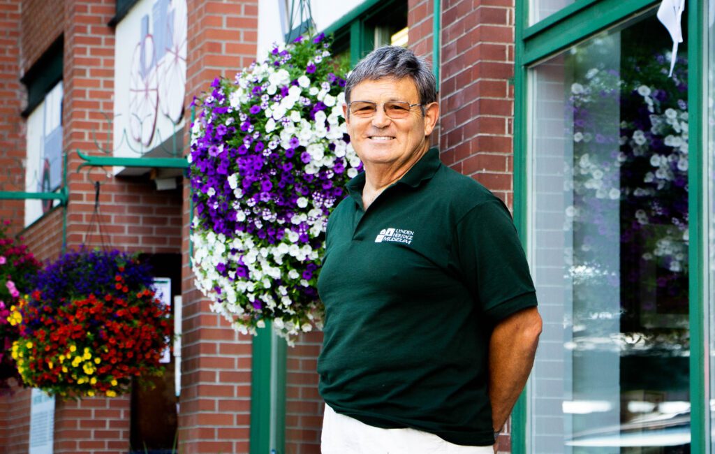 Surveyor historian Denny DeMeyer stands in front of the Lynden Heritage Museum lined up with flowers hanging above.