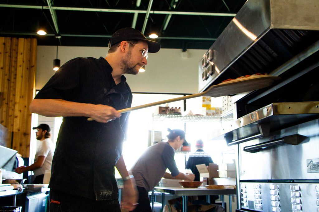 Ben Milne places a handmade pepperoni pizza into the oven as other staff work around him.