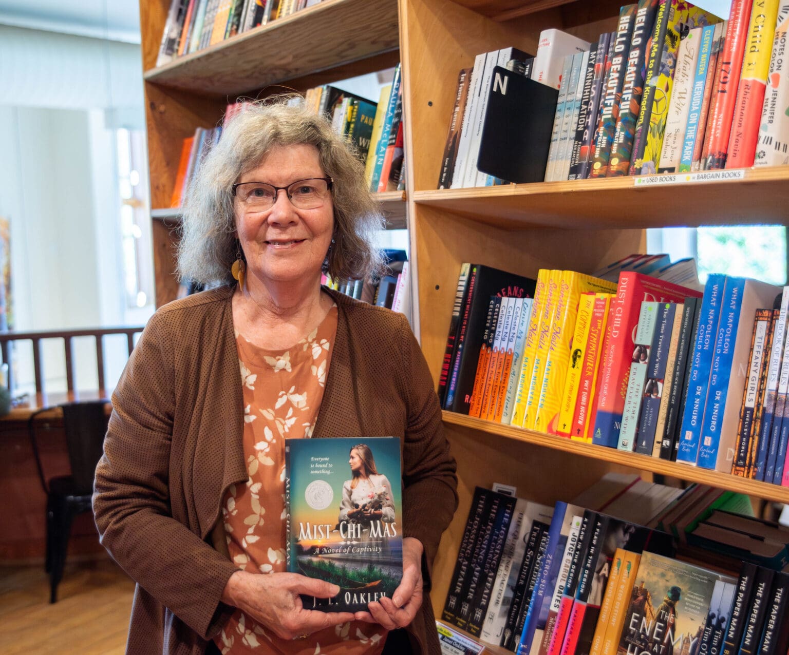 Janet Oakley holds a copy of her book "Mist-Chi-Mas" at Village Books in Fairhaven next to shelves of books stacks next to each other.