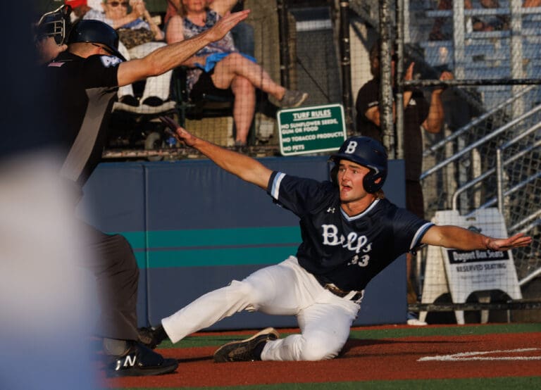 Sliding home, the Bellingham Bells' Coleman Schmidt spreads his arms in reaction as another person raises their arms to let people know of his ruling.