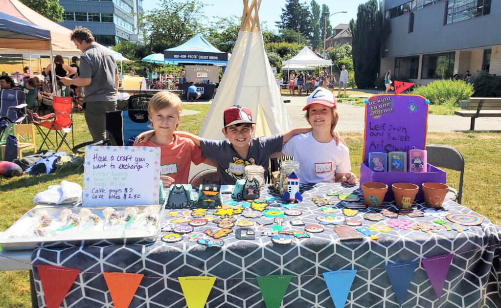 A booth set up by three kids smile for the camera as their table showcases various items for sale like cake pops and hand crafts.