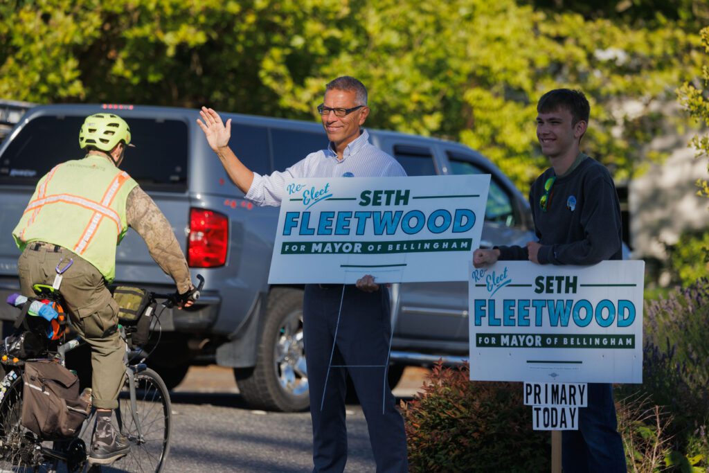 Mayor Seth Fleetwood, left, waves to drivers and bikers next to a younger man leaning on his sign.