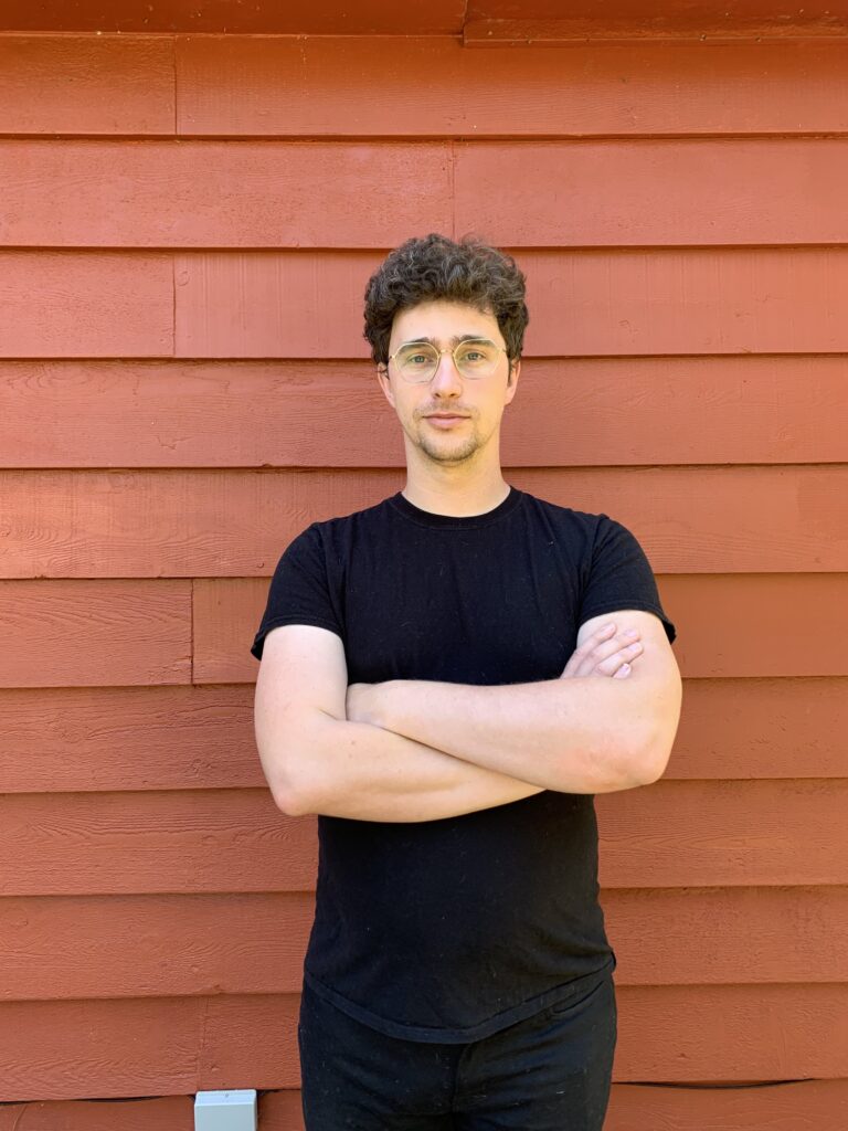 Victor Simpson poses for a photo in front of an orange wall with his arms crossed.