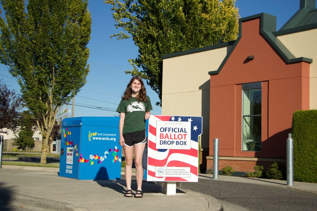 Hannah Nyland, 18, drops off her ballot as she smiles while leaning against the official ballot drop box.