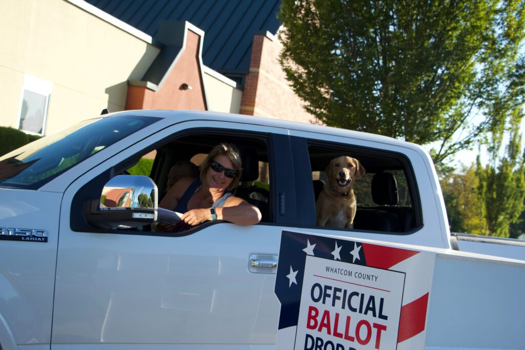 Teresa Kats and her dog, Rainer drives by the ballot box to drop off their ballot as Rainer looks at the camera in curiosity.
