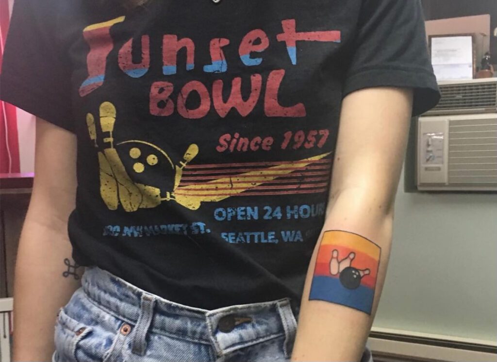 A young woman is wearing a t-shirt that says "Sunset Bowl Since 1957" after getting a tattoo on her forearm of three bowling pins and a ball with a sunset background.