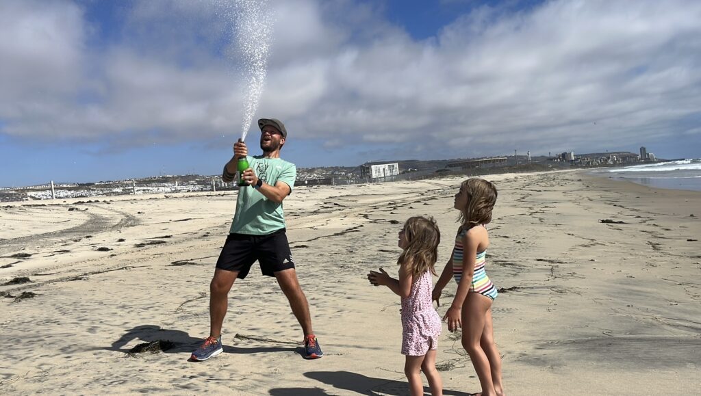 Marcus Nuckolls' daughters watch him as he pops a bottle of Champagne on the beach.