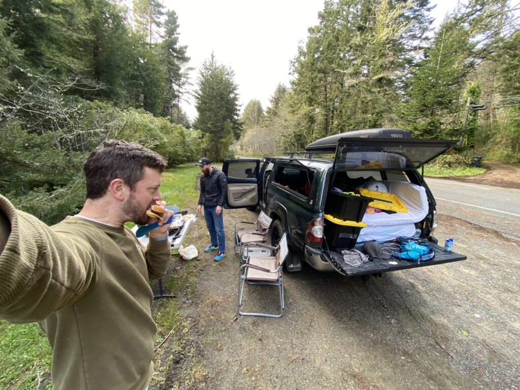 Marcus Nuckolls eats a burger on the side of the road as his friend, Troy Duvall, looks at the table of drinks and food.