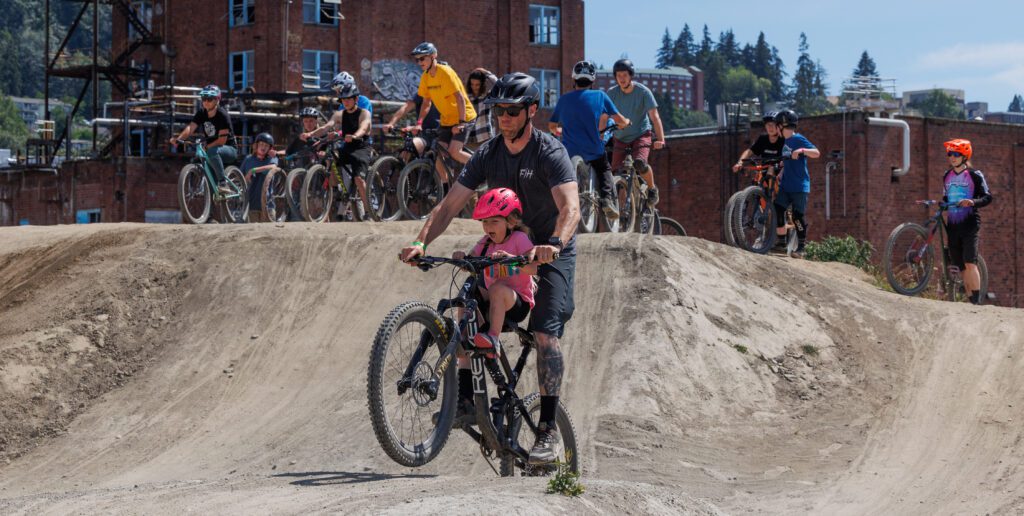 Kyle Spain rides a bicycle with his daughter, Hattie, as other cyclers wait their turn on the top of the sandy dune.