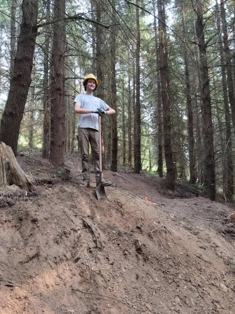 Aidan Ritchie stands atop a slope leaning on a shovel while wearing a yellow hard hat for safety.