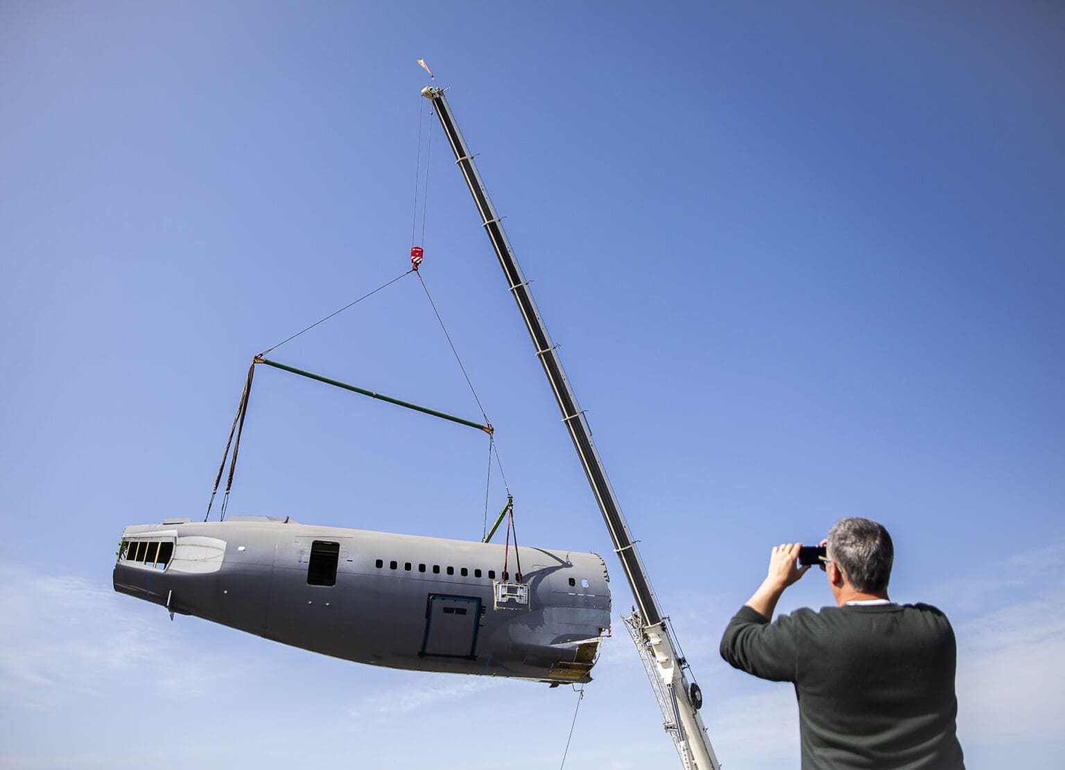 People gather and take photographs of a donated fuselage being moved by cranes.