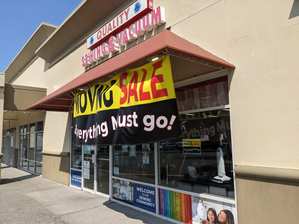 The storefront of Quality Sewing & Vacuum has a large banner hanging below the awning letting customers know of their moving sale.