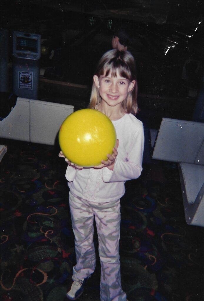 A girl smiles for the camera while holding a neon yellow bowling ball at a bowling alley.