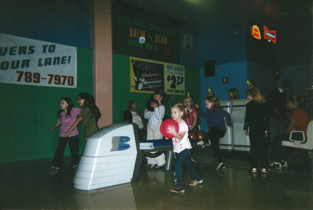 A young girl holding a pink bowling ball with two hands walks up to bowl as other children dance around her in party hats.