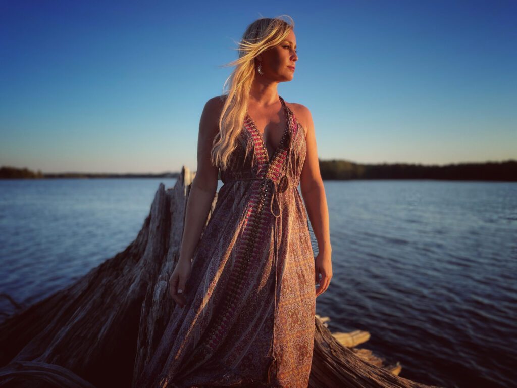 Sofia Talvik looks away from the camera as she stands on a large dead tree as the sun set colors shine on her.