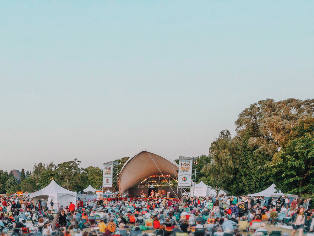 The Vancouver Folk Music Festival filled with visitors sitting on chairs or picnic mats as a live performers play music on stage.