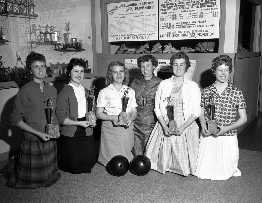 A black and white photo shows five girls in skirts with their bowling coach, posing for a photo as they hold bowling trophies.