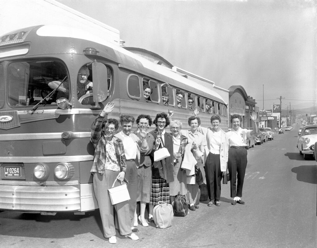 A black and white photograph shows a group of women standing outside an old bus.