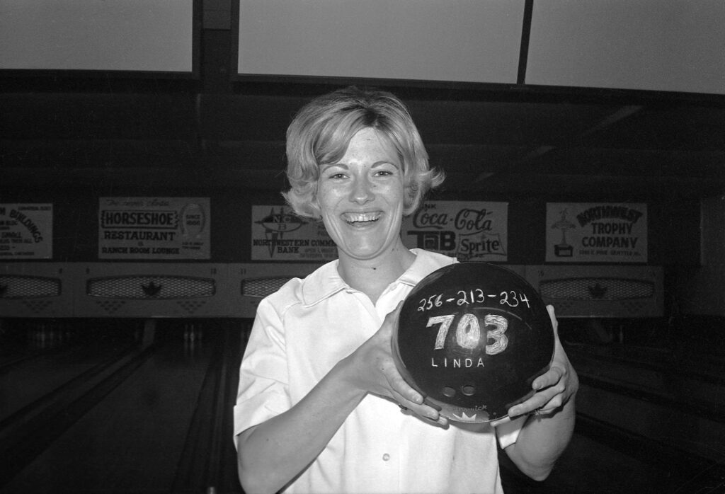 A black and white photograph shows a woman in a bowling alley holding a bowling ball that says "256-213-234, 703, Linda."