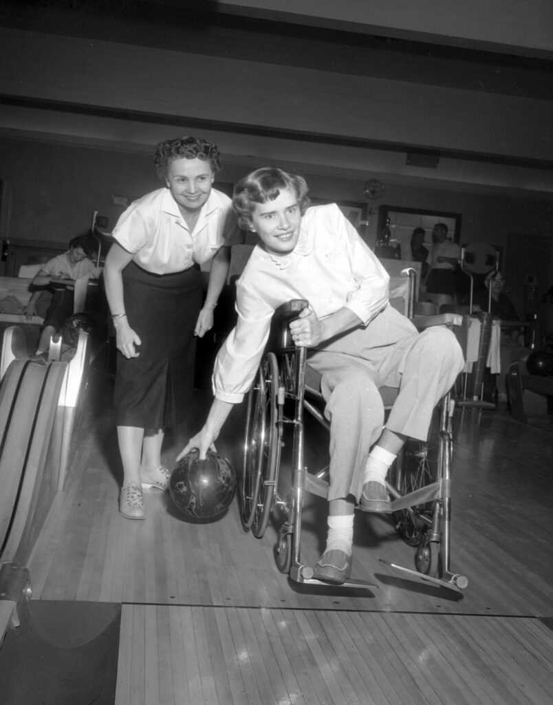 A black and white photograph shows a woman in a wheelchair bowling while another woman stands beside her.